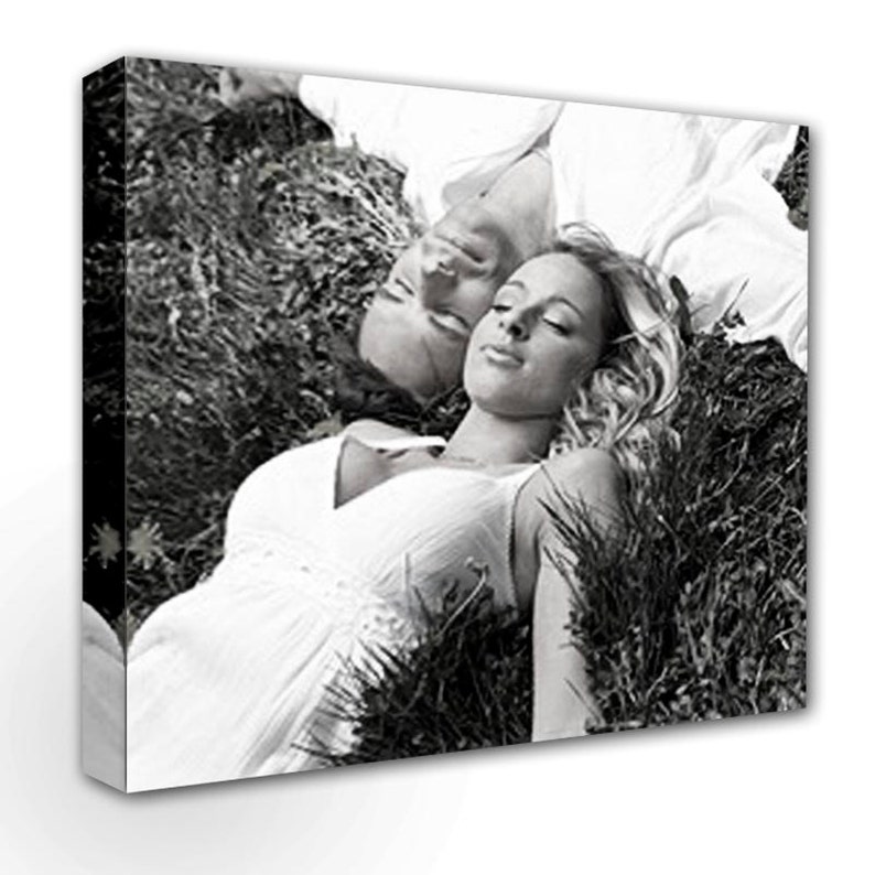 May include: A black and white photograph of a couple lying in a field of grass. The woman is wearing a white dress and has her eyes closed. The man is wearing a white shirt and has his eyes closed. The couple is lying close together, with their heads resting on each other's shoulders.