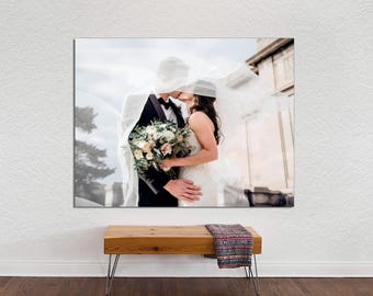 May include: A bride and groom kiss under a white veil, the bride is wearing a white dress and the groom is wearing a black suit. They are standing in front of a building with a white facade.