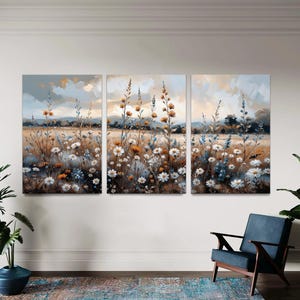 Op de afbeelding: Een drieluik canvas kunstset die een veld met wilde bloemen in een zachte, impressionistische stijl afbeeldt. Het kunstwerk toont witte, gele en blauwe bloemen tegen een achtergrond van een gedempte lucht en een gouden veld. Het kleurenpalet is zacht en natuurlijk.