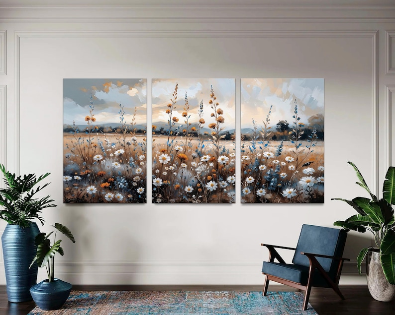 Op de afbeelding: Een drieluik canvas kunstset die een veld met wilde bloemen in een zachte, impressionistische stijl afbeeldt. Het kunstwerk toont witte, gele en blauwe bloemen tegen een achtergrond van een gedempte lucht en een gouden veld. Het kleurenpalet is zacht en natuurlijk.