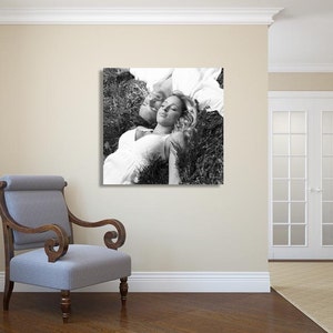 May include: A black and white photograph of a couple lying in a field of grass. The woman is wearing a white dress and has her eyes closed. The man is lying on his side with his head resting on her shoulder.