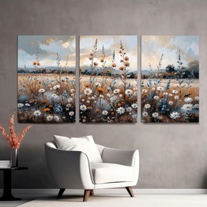 Op de afbeelding: Een driedelig canvas kunstwerk dat een veld met wilde bloemen in een impressionistische stijl toont. Het kunstwerk toont witte, gele en blauwe bloemen met een gedempt kleurenpalet, wat een serene landschap cre&euml;ert. Het kunstwerk wordt boven een witte fauteuil getoond.