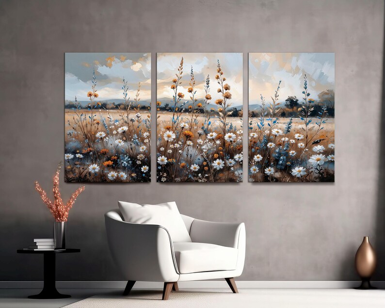 Op de afbeelding: Een driedelig canvas kunstwerk dat een veld met wilde bloemen in een impressionistische stijl toont. Het kunstwerk toont witte, gele en blauwe bloemen met een gedempt kleurenpalet, wat een serene landschap cre&euml;ert. Het kunstwerk wordt boven een witte fauteuil getoond.