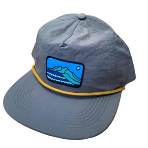 May include: A grey nylon cap with a flat peak and a yellow rope detail. The cap features a rectangular patch with a mountain and water scene in blue and teal. A small grey button is on top.