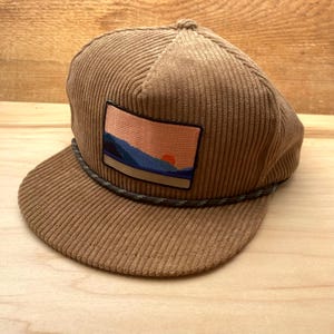 May include: A brown corduroy baseball cap with a patch featuring a sunset over mountains. The cap has a green and grey rope detail around the brim.