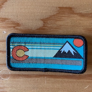 May include: Rectangular patch with a Colorado state flag design. It features a mountain range, a setting sun, and the state's 'C' logo in red, yellow, and white. The background has horizontal stripes in shades of blue, grey, and teal.