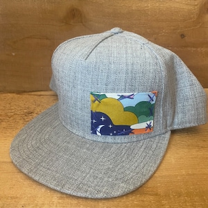 May include: A grey, flat-brimmed baseball cap with a colourful, stitched patch on the front. The patch features a design of clouds, stars, and a crescent moon.