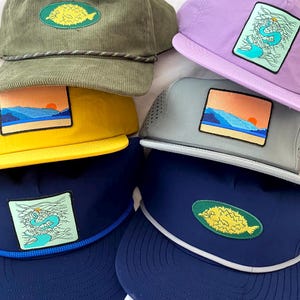 Könnte beinhalten: Fünf verschiedene Baseballkappen mit verschiedenen gestickten Designs. Die Kappen sind in verschiedenen Farben, darunter Grün, Lila, Gelb, Grau und Blau. Die Designs beinhalten einen Fisch, eine Berglandschaft und eine Welle.