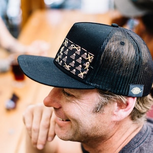 May include: A black and gold patterned trucker hat with a mesh back. The hat has a small gold fish logo on the side.