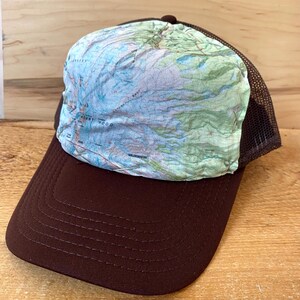 May include: A brown and black mesh trucker hat with a topographic map design on the front. The map shows the area around Mount Wood.