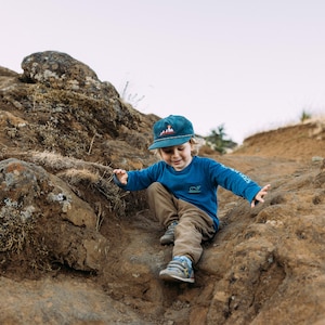 May include: A young child wearing a blue long-sleeved shirt and a blue and red baseball cap is climbing on rocks. The child is looking down at the rocks.