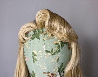 May include: A blonde wig with a large, curled section on top. The wig is styled in a vintage updo and is displayed on a mannequin head with a floral pattern.