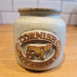 May include: A stoneware jar with a light beige speckled finish. The jar features a brown embossed design with the words "CORNISH GOODIES" and a cow illustration. The jar has a slightly flared rim and a cylindrical shape.