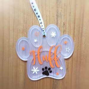 May include: A clear plastic paw print ornament with the name "Fluff" written in orange. The ornament is decorated with white snowflakes and stars. A black paw print is in the center of the ornament.