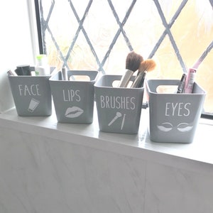 May include: Four grey plastic containers with white labels and icons. The containers are labelled "Face", "Lips", "Brushes", and "Eyes". The containers are filled with makeup brushes and other makeup items.
