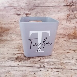 May include: A light grey square storage container featuring the name "Taylor" and the letter "T" in white. The container has a rectangular opening and a wooden handle. The background is a light brown wooden surface.