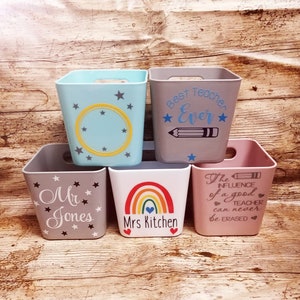 May include: Five square storage bins with different designs. The bins are light blue, grey, white, and pink. The designs include stars, a rainbow, a pencil, and text that says "Best Teacher Ever", "Mr. Jones", "Mrs. Kitchen", and "The Influence of a good Teacher can never be Erased".