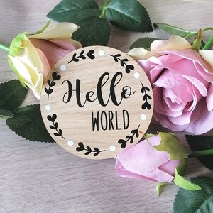 May include: A round wooden sign with a white background and black lettering. The sign says "Hello World" with a decorative border of leaves and white dots.