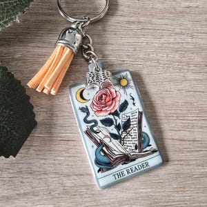 May include: A rectangular keychain with a tarot card design. The card shows a pink rose, a snake, and an open book, with the words "THE READER" at the bottom. It has a silver tassel and a silver key ring.