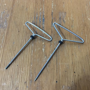 May include: Two metal T-shaped tools with a pointed end and a wire handle. Each tool has a small black rubber ring near the handle. The tools are silver and appear to be made of metal wire.