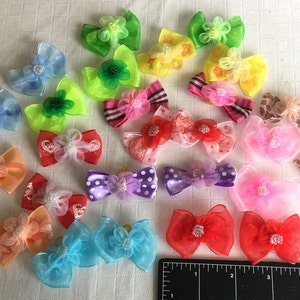 May include: A collection of colorful hair bows made of sheer fabric with various embellishments. The bows are arranged on a white surface with a ruler in the background.