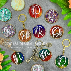 May include: A set of 10 resin keychains with different letters of the alphabet, each with a gold glitter background and a white floral design. The keychains are arranged in a circle on a gray surface with green foliage.