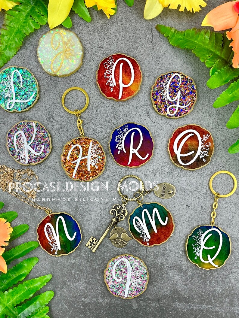 May include: A set of 10 resin keychains with different letters of the alphabet, each with a gold glitter background and a white floral design. The keychains are arranged in a circle on a gray surface with green foliage.
