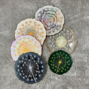 May include: Six colorful coasters with a dandelion design. The coasters are made of resin and have a glossy finish. The coasters are in various colors, including pink, green, black, and white.
