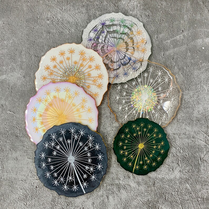 May include: Six colorful coasters with a dandelion design. The coasters are made of resin and have a glossy finish. The coasters are in various colors, including pink, green, black, and white.