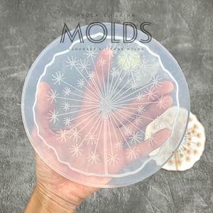 May include: A clear silicone mold with a raised design of a dandelion. The mold is round and has a scalloped edge. The mold is held in a hand.