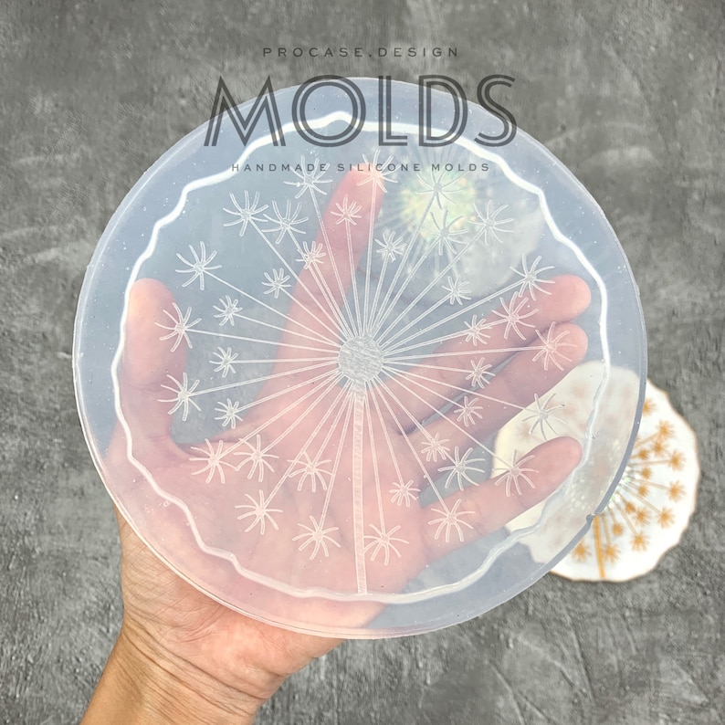 May include: A clear silicone mold with a raised design of a dandelion. The mold is round and has a scalloped edge. The mold is held in a hand.