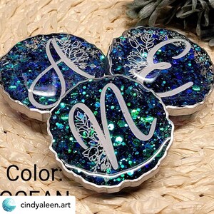 May include: Three round resin ornaments with a silver rim. Each ornament features a different letter, A, E, and N, in a white cursive font with a floral design. The ornaments are filled with blue and green glitter.