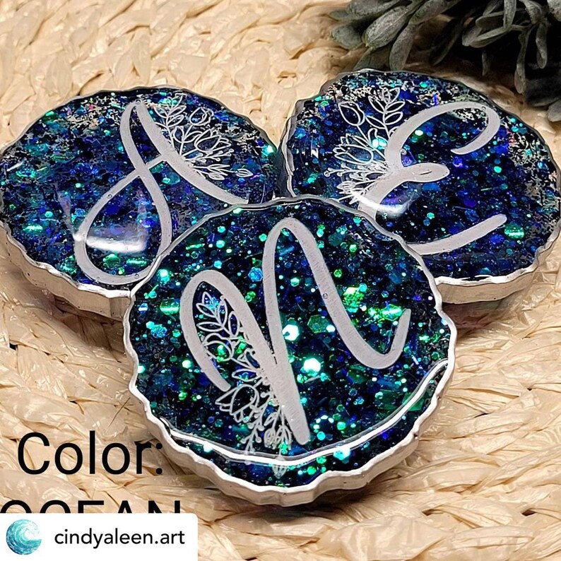 May include: Three round resin ornaments with a silver rim. Each ornament features a different letter, A, E, and N, in a white cursive font with a floral design. The ornaments are filled with blue and green glitter.