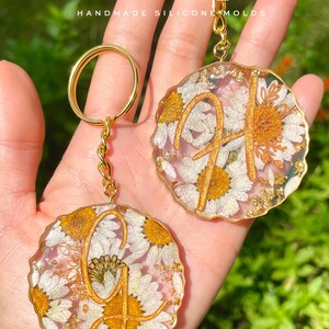 May include: Two gold keychains with round resin charms featuring pressed white flowers and gold glitter. The charms have a gold letter "G" and "H" in the center. The text "PROCASE.DESIGN.MOLDS HANDMADE SILICONE MOLDS" is visible in the image.
