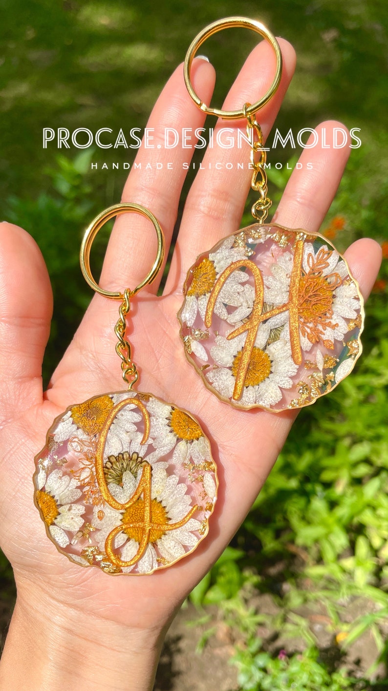 May include: Two gold keychains with round resin charms featuring pressed white flowers and gold glitter. The charms have a gold letter "G" and "H" in the center. The text "PROCASE.DESIGN.MOLDS HANDMADE SILICONE MOLDS" is visible in the image.