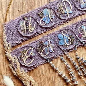 May include: A purple burlap banner with the words "Happy Easter" spelled out in gold lettering on individual resin circles. Each circle is filled with pressed flowers and leaves.