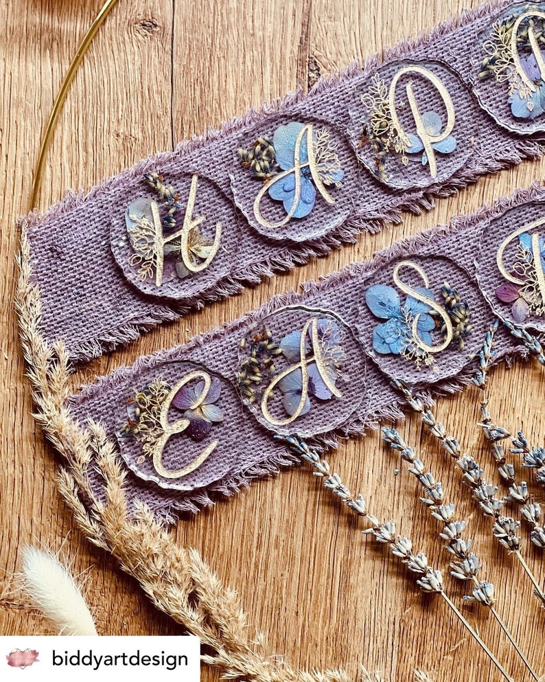 May include: A purple burlap banner with the words "Happy Easter" spelled out in gold lettering on individual resin circles. Each circle is filled with pressed flowers and leaves.