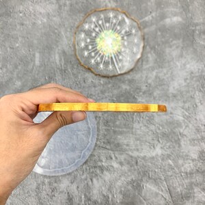 May include: A close-up of a round, wooden coaster with a light brown color. The coaster is being held by a hand, and the edge of the coaster is visible.
