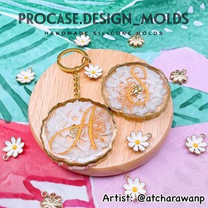 May include: Two gold-rimmed resin keychains with white floral designs and gold lettering. The keychains are on a wooden surface with white daisies scattered around. The text "PROCASE.DESIGN_MOLDS" and "HANDMADE SILICONE MOLDS" is at the top of the image. The text "Artist: @atcharawanp" is at the bottom of the image.
