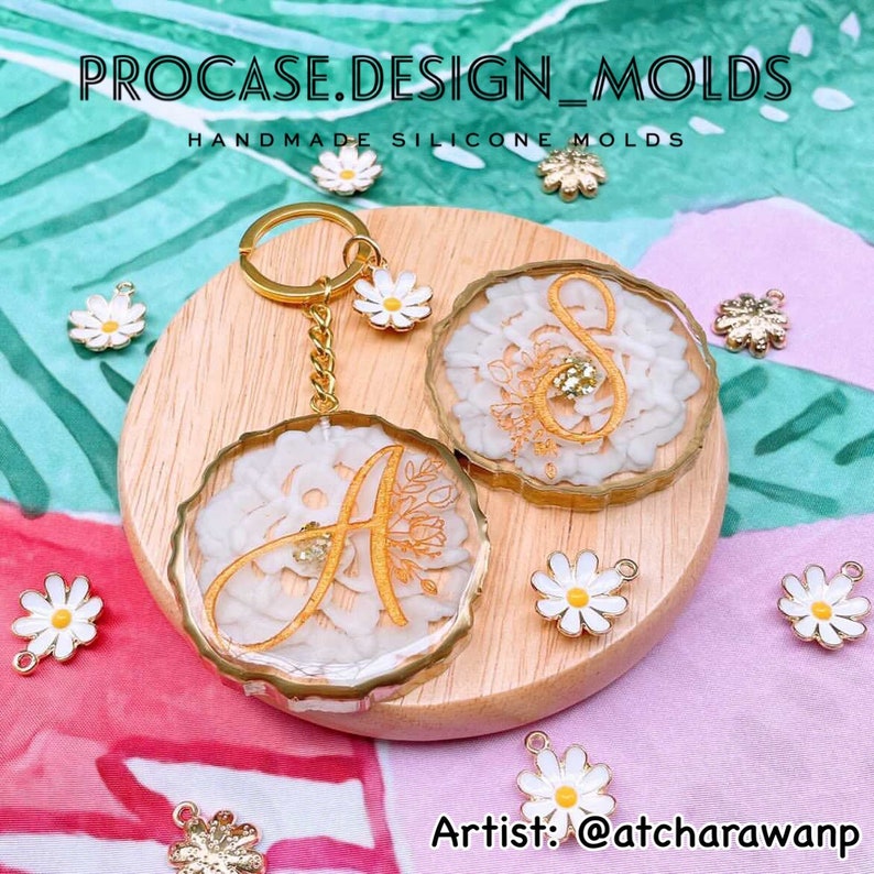 May include: Two gold-rimmed resin keychains with white floral designs and gold lettering. The keychains are on a wooden surface with white daisies scattered around. The text "PROCASE.DESIGN_MOLDS" and "HANDMADE SILICONE MOLDS" is at the top of the image. The text "Artist: @atcharawanp" is at the bottom of the image.