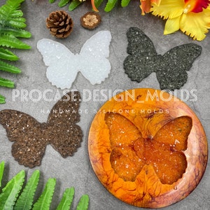 May include: Four butterfly-shaped silicone molds, two with glitter embedded in the resin, one with brown glitter and one with black glitter. The fourth mold is a round mold with a butterfly cutout and orange glitter embedded in the resin. The molds are on a gray surface with green foliage and a small pine cone.