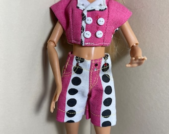 May include: A pink and white polka dot crop top and shorts outfit for a doll. The top has a white collar and four white buttons. The shorts have a black and white polka dot pattern on a white background.