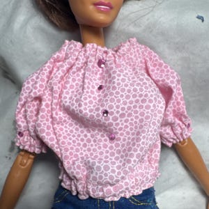 May include: A pink and white polka dot blouse with a ruffled neckline and short sleeves. The blouse has pink rhinestones down the front.