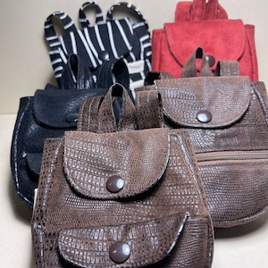 May include: Five miniature backpacks in different colors and materials. The backpacks are made of black and white striped fabric, black leather, brown leather with a textured pattern, and red suede.