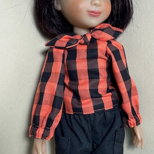 May include: A doll wearing a red and black plaid shirt with a bow tie and black pants with pockets.