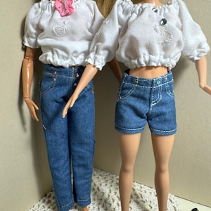 May include: Two dolls wearing white off-the-shoulder tops with embroidered details and denim pants and shorts. The doll on the left is wearing long denim pants with rhinestones, while the doll on the right is wearing denim shorts.