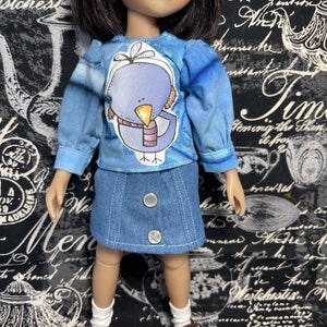 May include: A doll wearing a blue and white long-sleeved shirt with a cartoon bird graphic. The doll is also wearing a denim skirt and brown shoes.