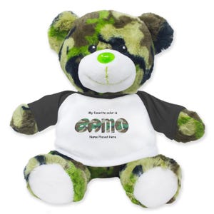 May include: A plush teddy bear with a camouflage pattern in shades of green, brown, and black. The bear wears a white shirt with black sleeves. The shirt has the text "My favorite color is camo" and "Name Placed Here". The bear has a green nose.