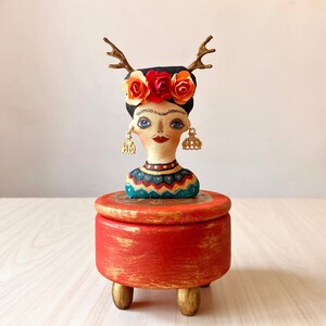 May include: A red wooden box with gold accents and a painted figure of a woman with a flower crown and antlers on top. The woman is wearing a blue and green patterned shirt.