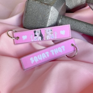Puede incluir: Dos llaveros rosas con un corazón blanco y un gráfico blanco de dos chicas de anime. Un llavero dice "SQUAT THAT" en letras blancas que gotean.